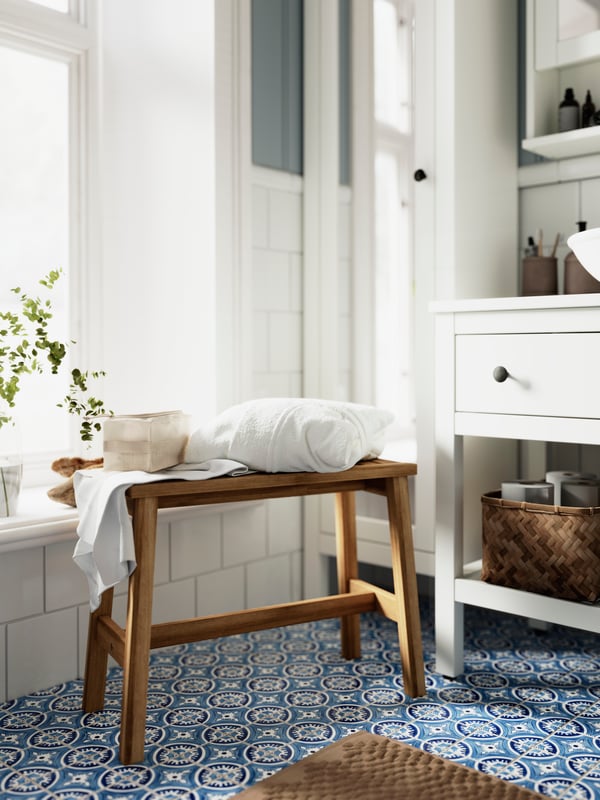 A bright, white-tiled bathroom with a blue mosaic floor has white HEMNES bathroom furniture and a wooden STOREDAMM bench.