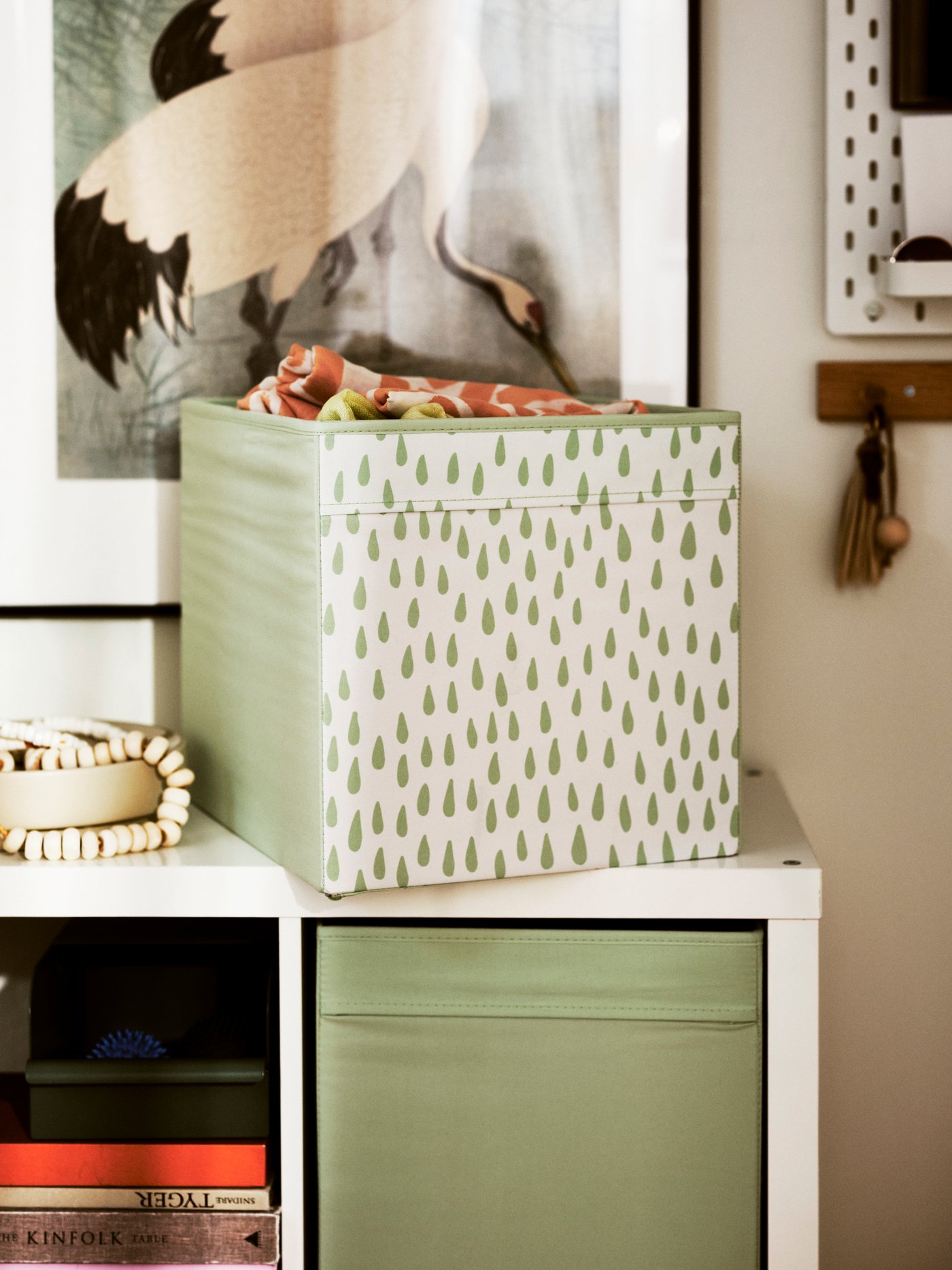 Une étagère bien rangée avec en haut une boîte de rangement vert et blanc à motif d’arbres, en dessous des compartiments contenant un bac vert et divers objets, le tout contre un mur décoré d’un oiseau encadré et d’un petit crochet en bois.