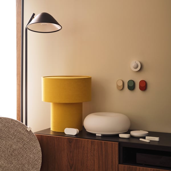 A MYGGSPRAY wireless motion sensor and three BILRESA remote controls are mounted on a wall above a TV bench next to a lamp.