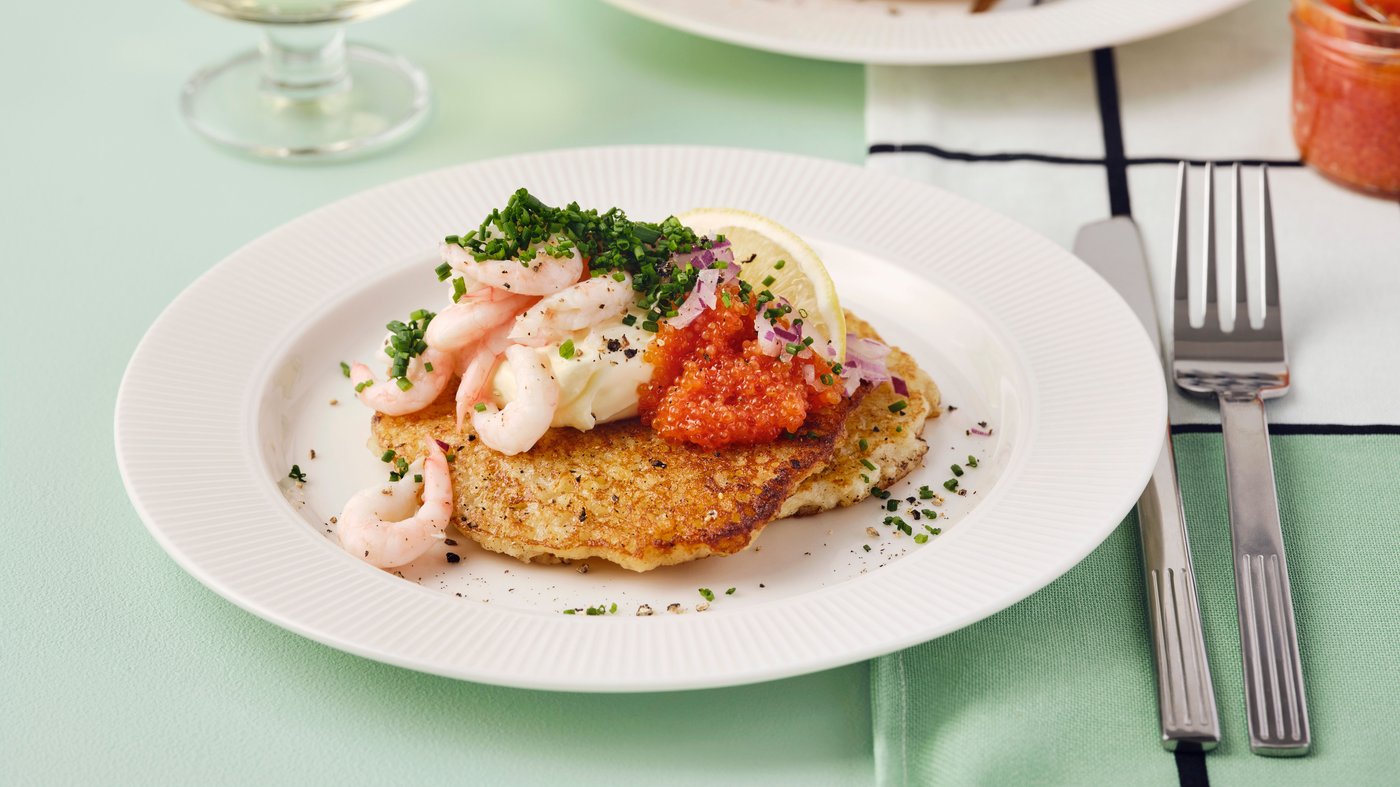 Dva zlatnosmeđa RÖSTI-ja sa škampima, kavijarom i kriškom limuna nalazi se na belom tanjiru na zelenom stolnjaku.