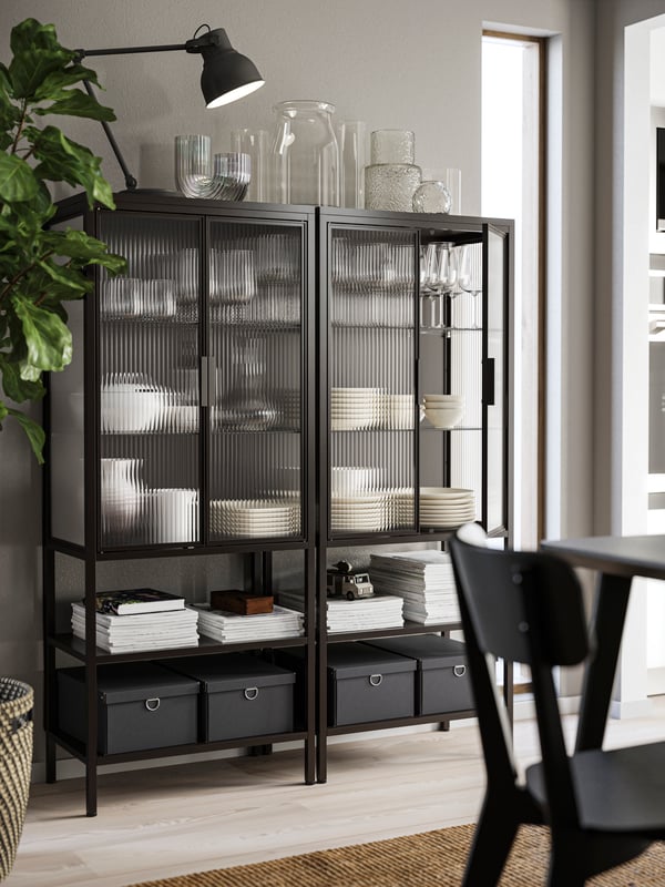 Two MOSSJÖN glass-door cabinets in anthracite have plates and glasses inside, vases on top and boxes on the lower shelves.