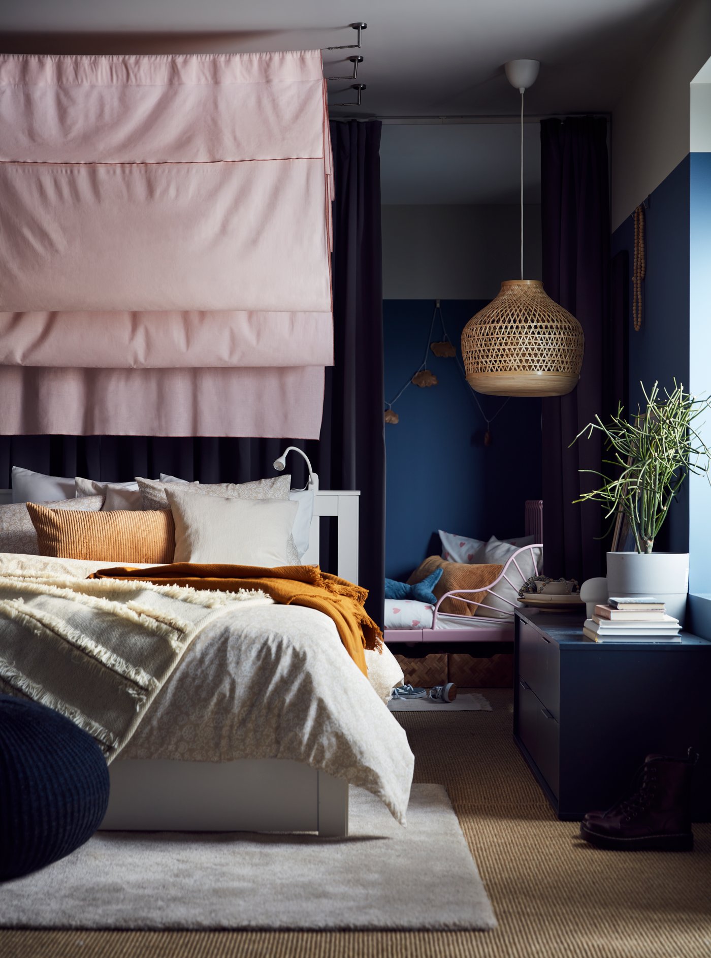 A white SONGESAND bed covered with light grey bed linen with a yellow OMTÄNKSAM throw on it and a pink curtain behind it.