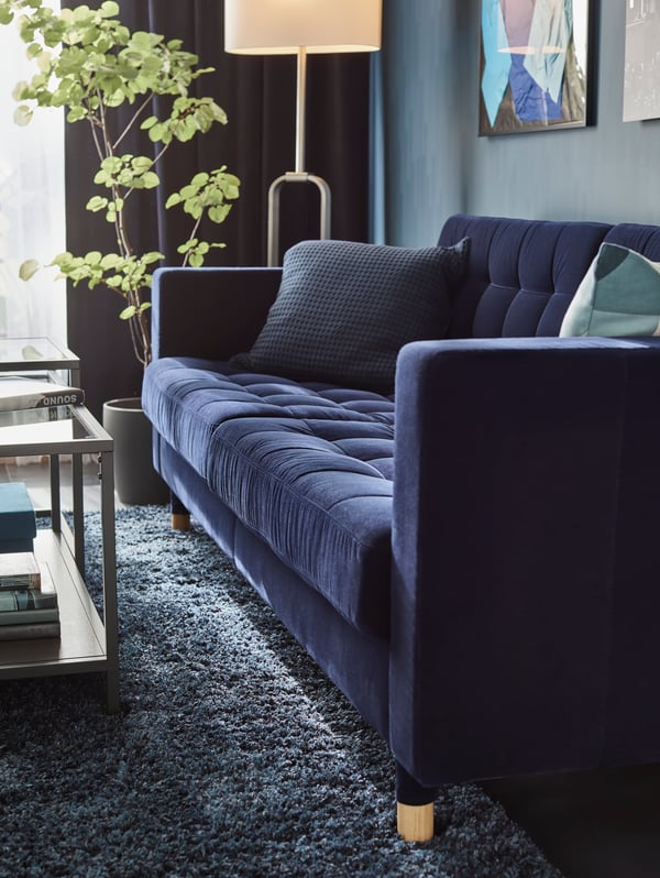A blue LANDSKRONA sofa with a wall of pictures behind it, a plant and floor lamp at one end and two glass tables in front.