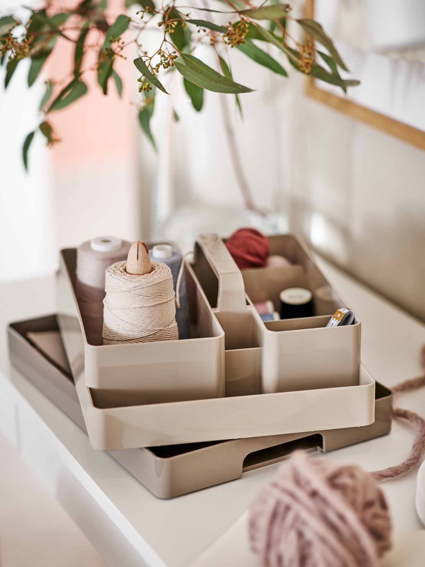 A grey-beige KUGGIS desk organiser set on a white surface holding different coloured sewing threads, with a plant overhead.
