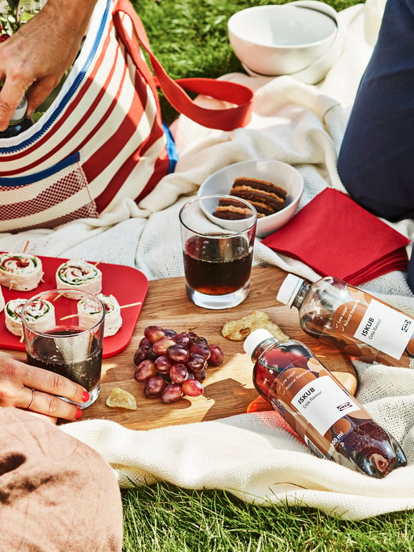 A blanket laid on the ground for a picnic, with bottles of ISKUB carbonated soft drink, napkins and diverse food items.