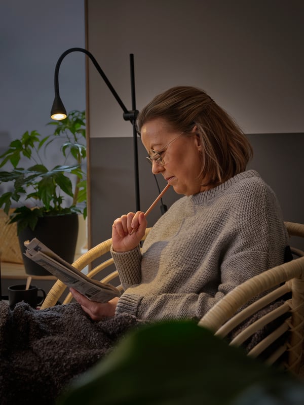 Light from a NÄVLINGE floor lamp helps a woman focus on finding clues for a puzzle as she sits in a BUSKBO armchair.