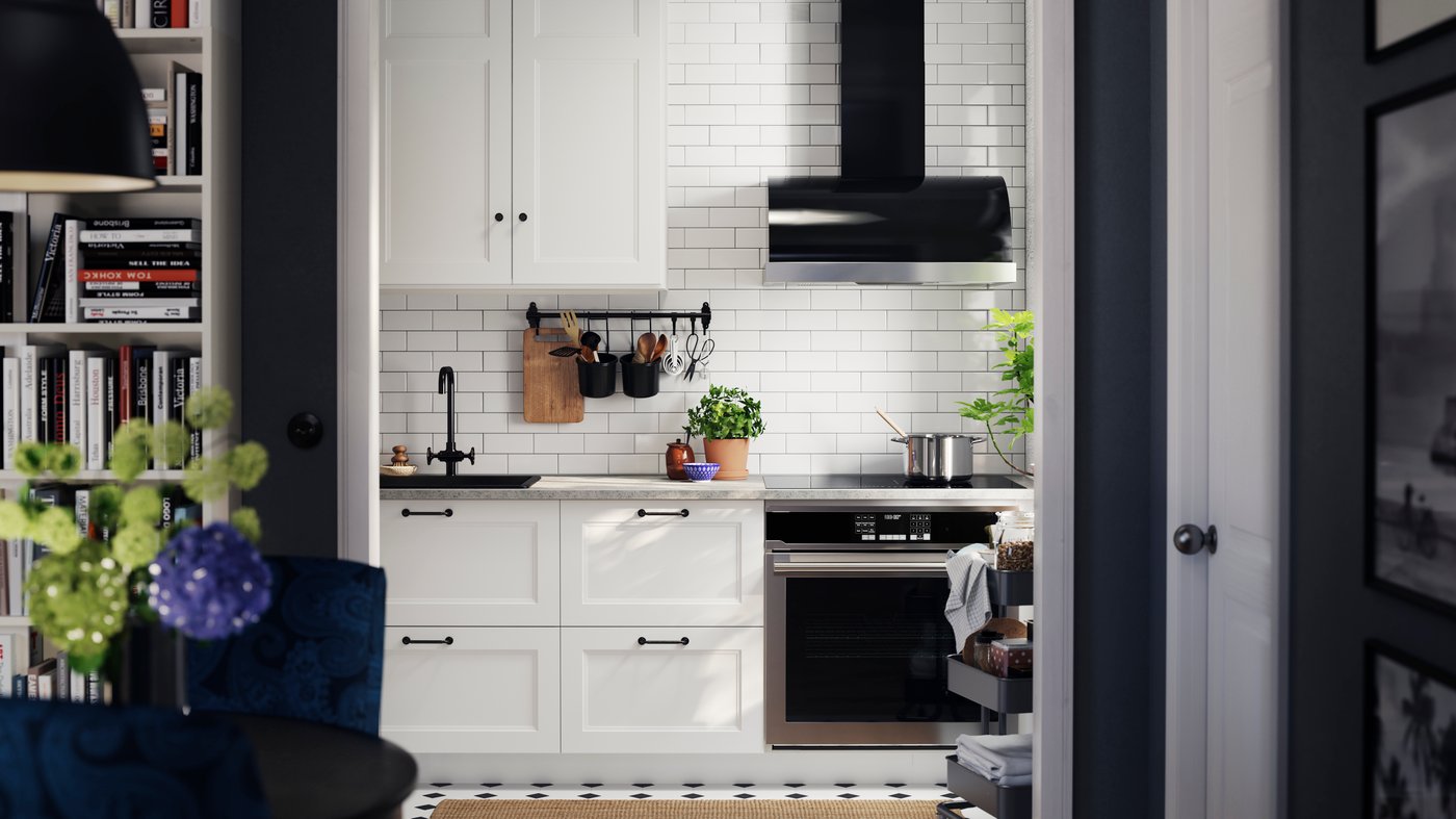 A kitchen with AXSTAD matte white kitchen fronts with black handles, a black tap, and potted plants, against tiled walls.