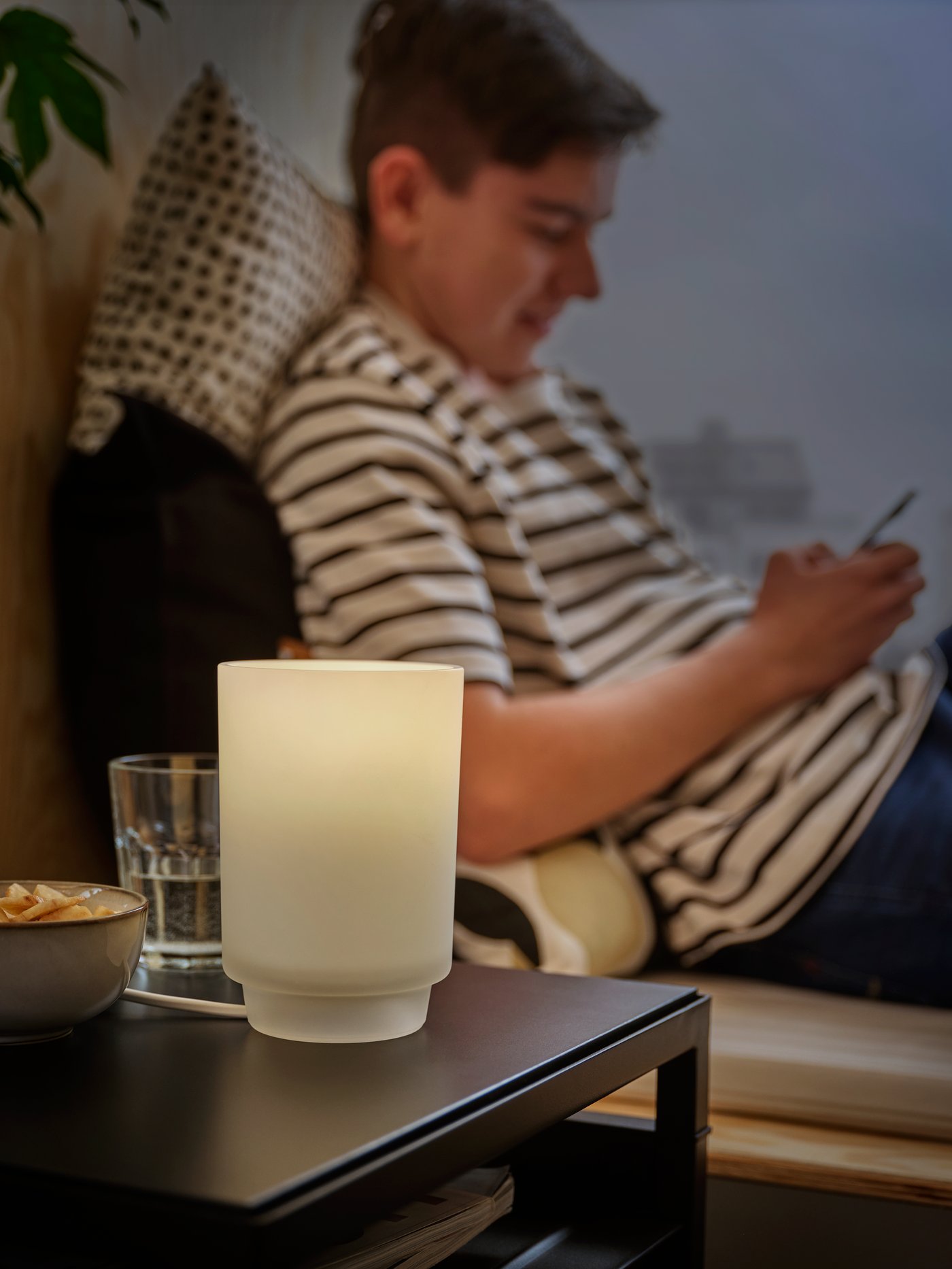 A frosted glass ISBRYTARE table lamp casts a cozy glow as a teenage boy in a striped shirt messages friends on his phone.