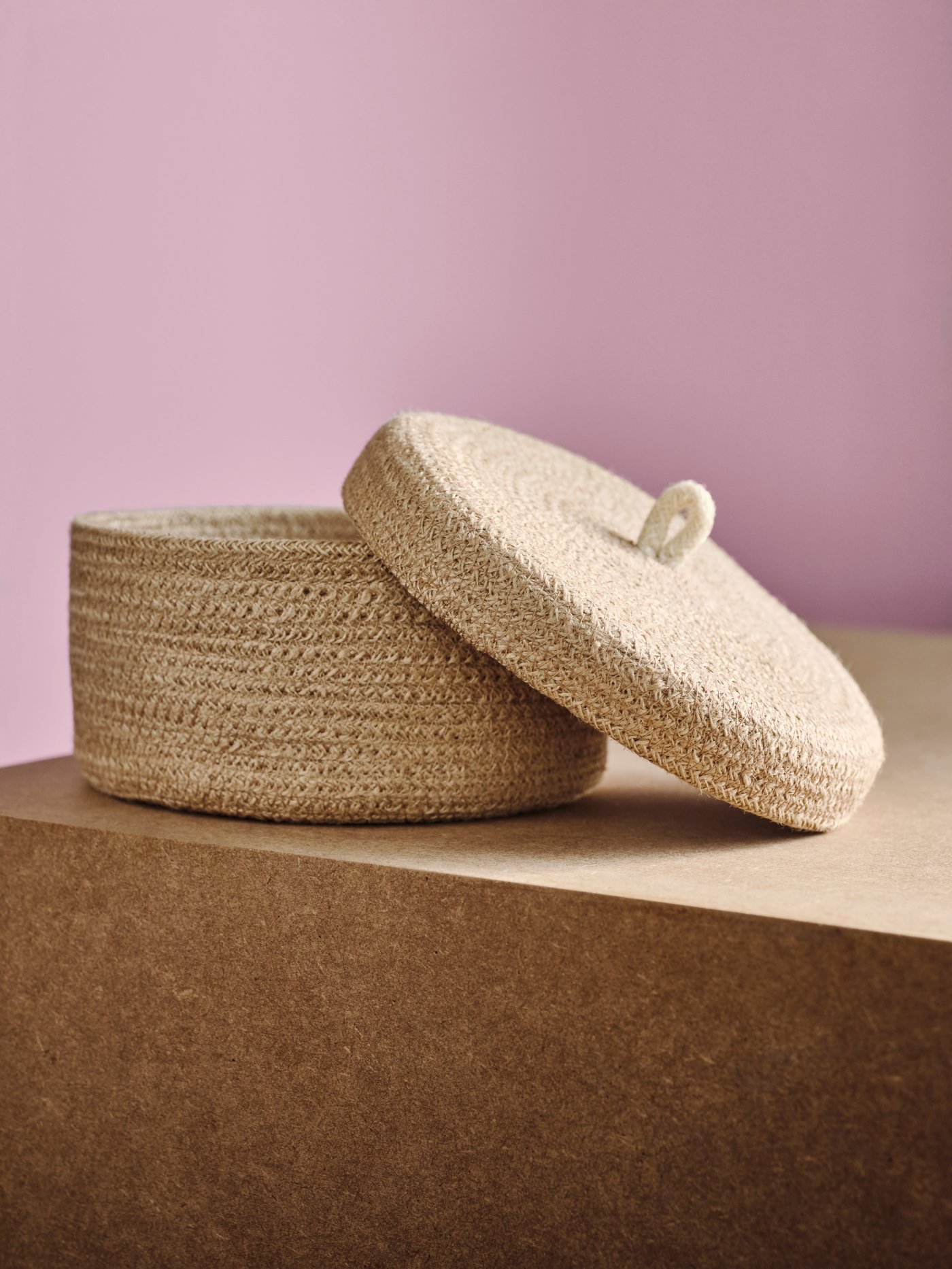 A LJUNGAN basket, made from jute, with lid is on top of brown stand.