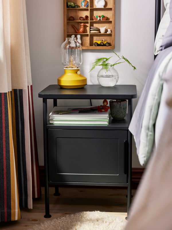 Close-up of GRÅFJÄLLET bedside table in anthracite with yellow TÄRNABY table lamp and small glass vase on top.