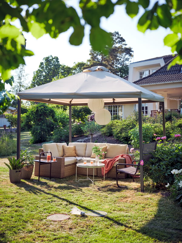 A SOLLERÖN corner outdoor sofa with beige cushions and a black armchair under a HIMMELSÖ gazebo on a sunny garden lawn.