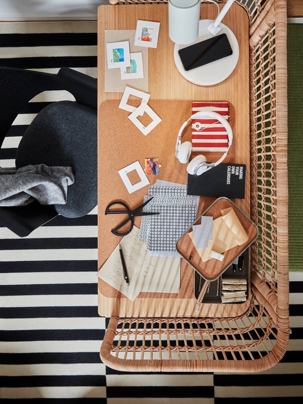 Auf einem FRYKSÅS Schreibtisch aus Rattan, der auf einem gestreiften STOCKHOLM Teppich steht, sind Zubehör, HARVMATTA Boxen und eine SUSIG Schreibunterlage aus Kork zu sehen.