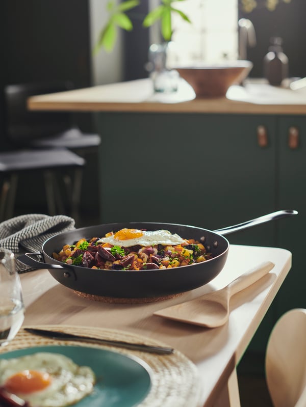 Une poêle à frire VARDAGEN avec du pyttipanna et une spatule à wok en hêtre sur une table en bouleau mise avec des assiettes FÄRGKLAR turquoise clair.