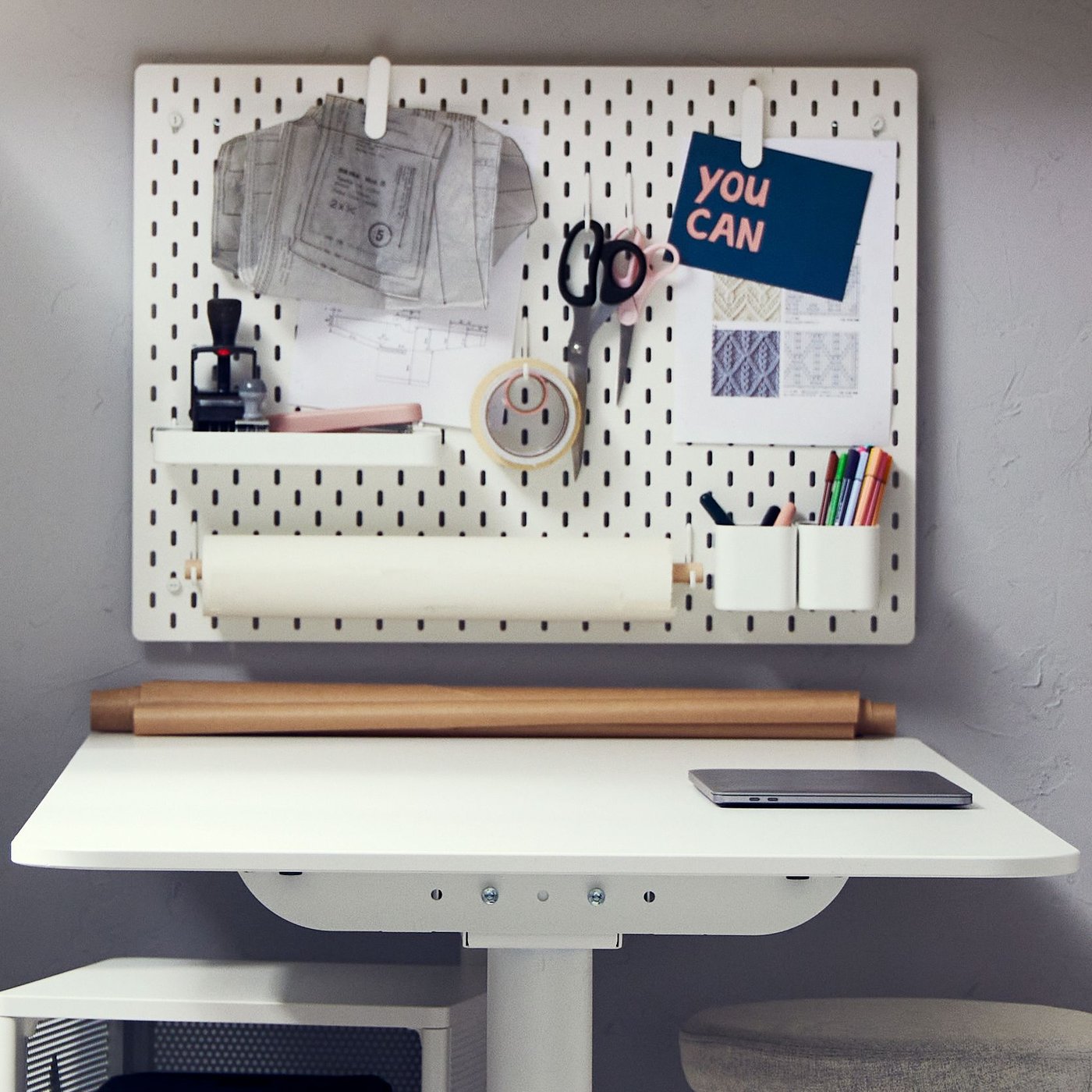 A white shelving unit with yarn, a light green armchair, a white desk with sit/stand support, pine cabinets and a pendant.
