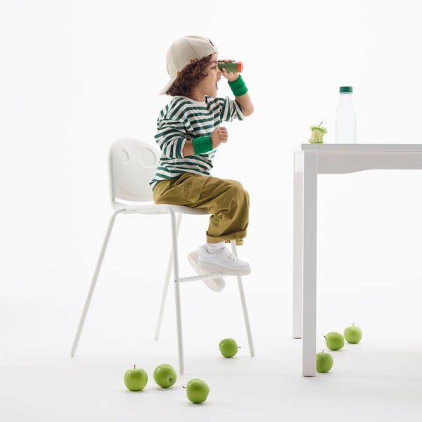 A 5-year-old is sitting in a white VILDGÅS junior dining chair, looking out over a dining table with a pair of binoculars.
