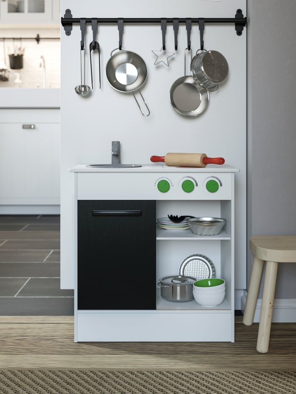 A white play kitchen with sliding door, toy kitchen utensils hanging on a rail, and a toy baking set.