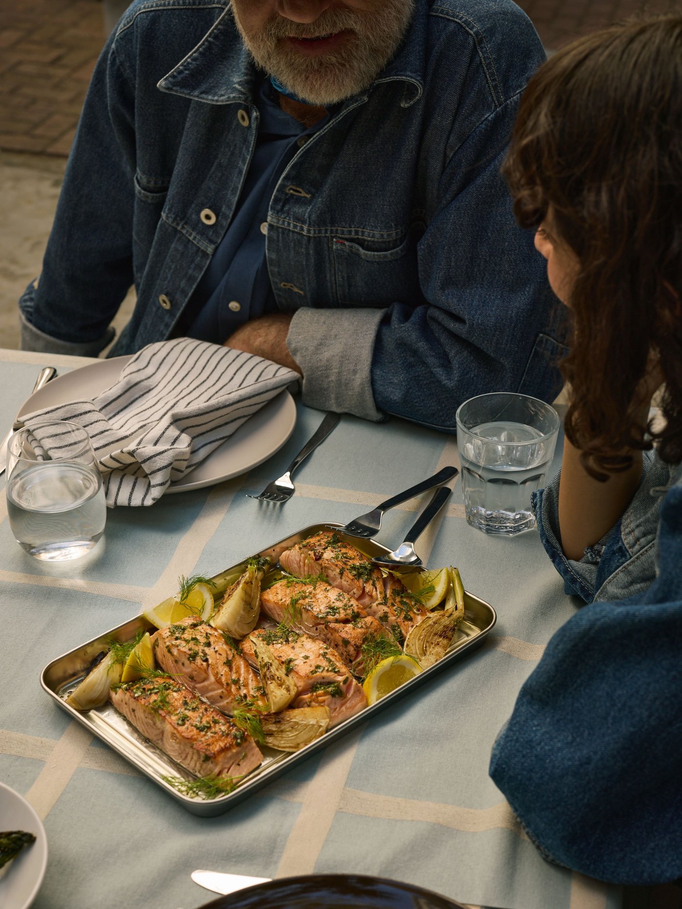Two people in jeans jackets sitting at a made dinner table, waiting to eat grilled food on a GRILLTIDER serving tray.