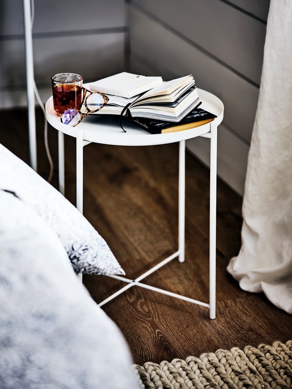 Une table basse GLADOM blanche dans le coin d’une chambre à côté d’un lit, avec des livres, des lunettes et une boisson dessus.