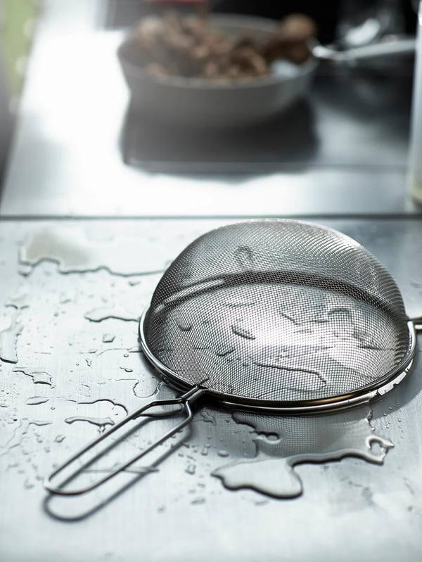 An IDEALISK strainer is placed on top of a wet stainless steel surface. In the background, there’s grilled food.