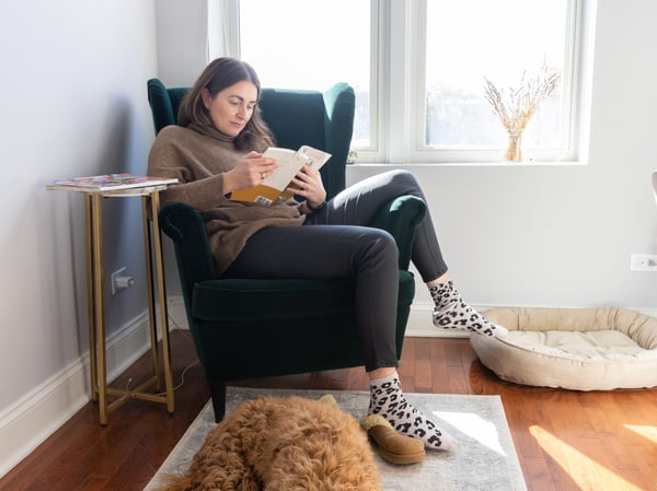 Lady sitting in sofa reading a book with her dog on the floor. 