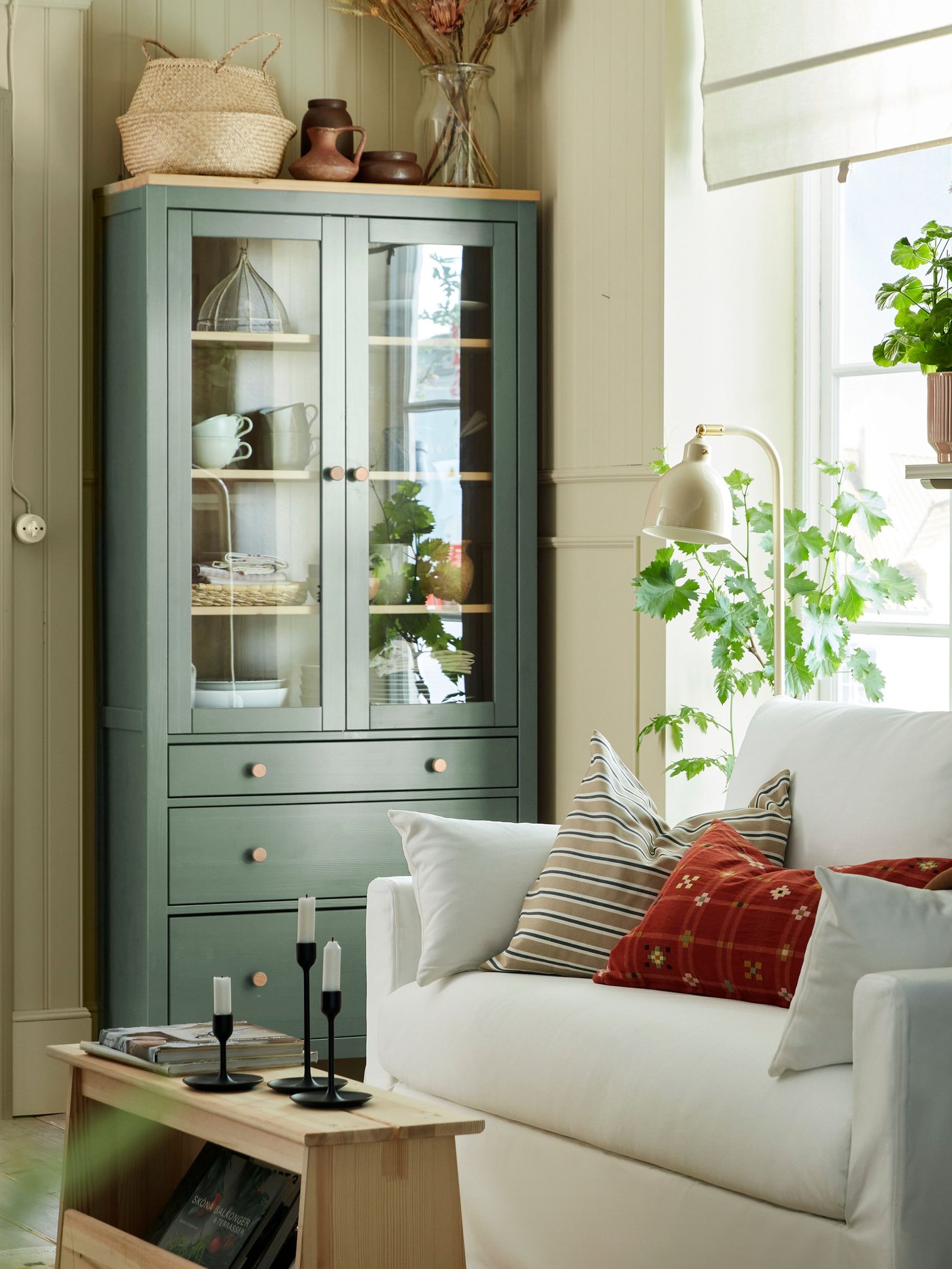Ein grau-grüner HEMNES Wohnzimmerschrank Mit Glastüren und Schubladen steht neben einem weissen Sofa am Fenster.