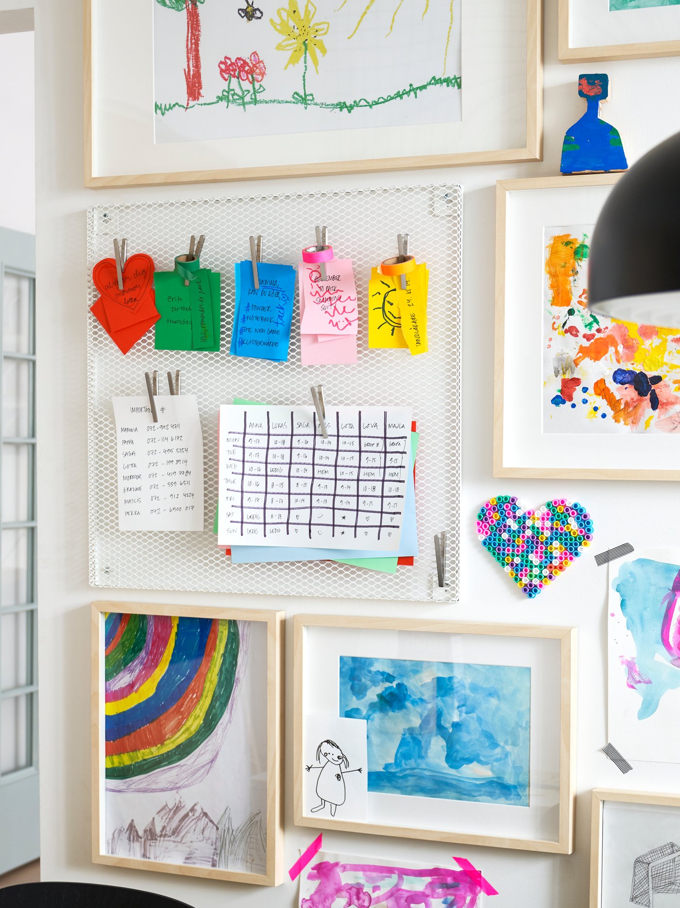 White wall with white memo board with magnets, colourful notes, calendars and daily organisers, wooden frames with drawings.