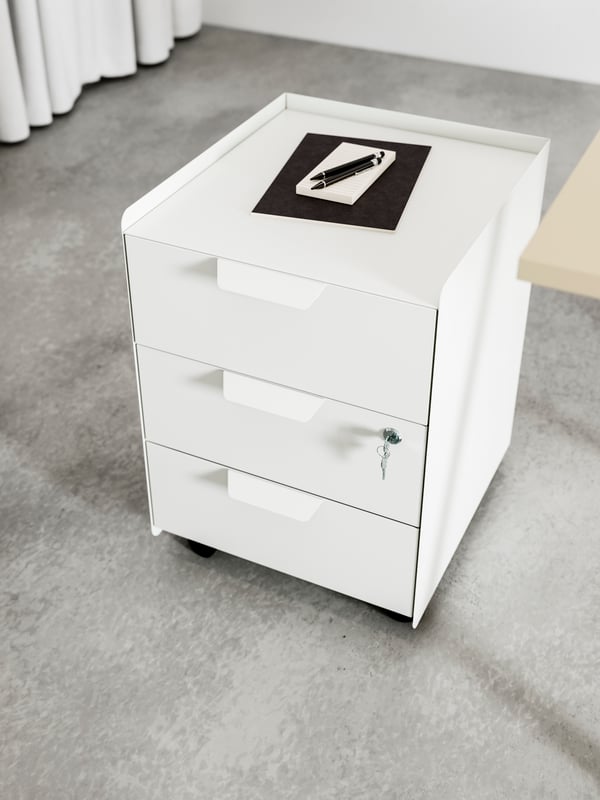 Next to a bed stands a white table with work accessories scattered on it and an TROTTEN drawer unit placed underneath.