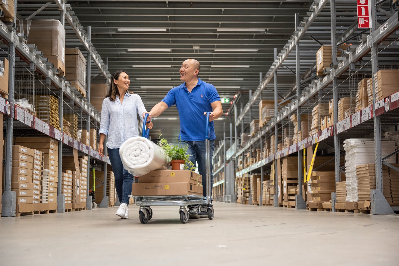 Two people walk through the IKEA self-serve warehouse, pushing a trolley with flat-packed boxes, a rolled mattress and a potted plant.