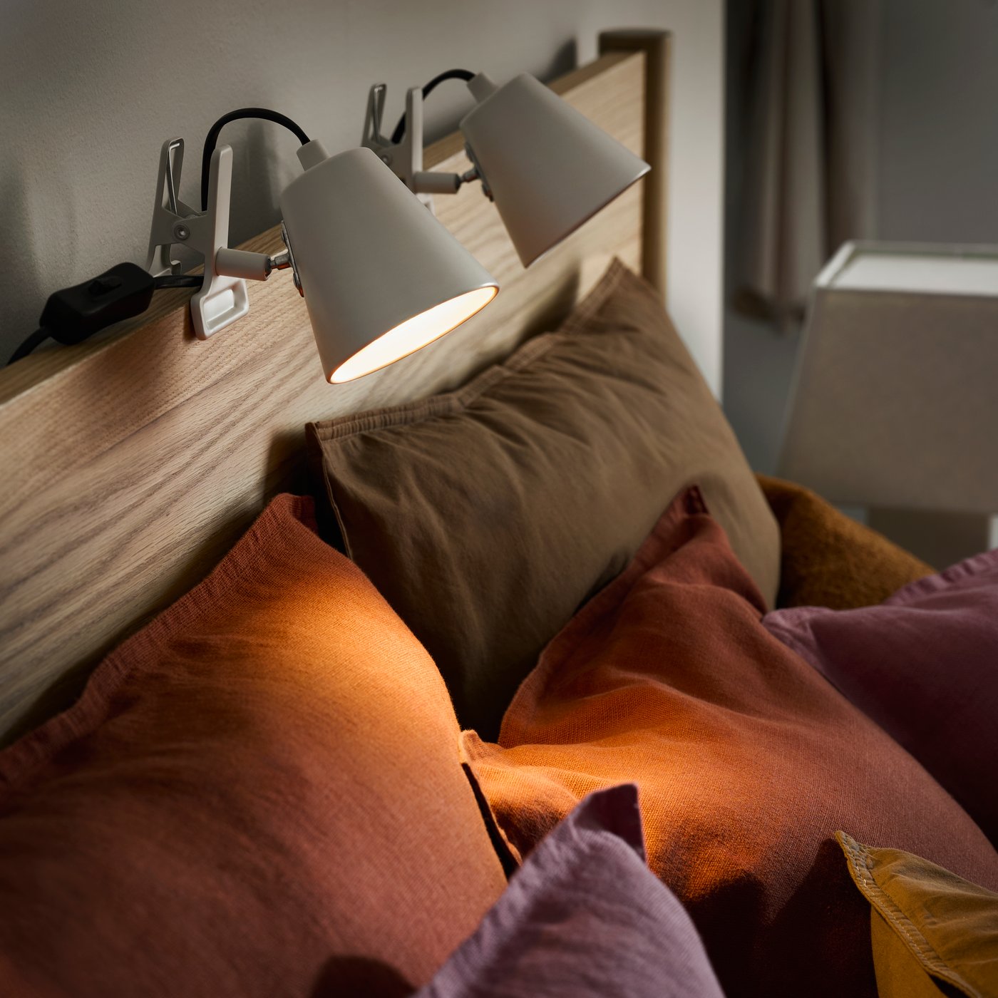A close up of HAVSSALLAD clamp stptlight on a light wooden bed frame with pink and brown pillows below.