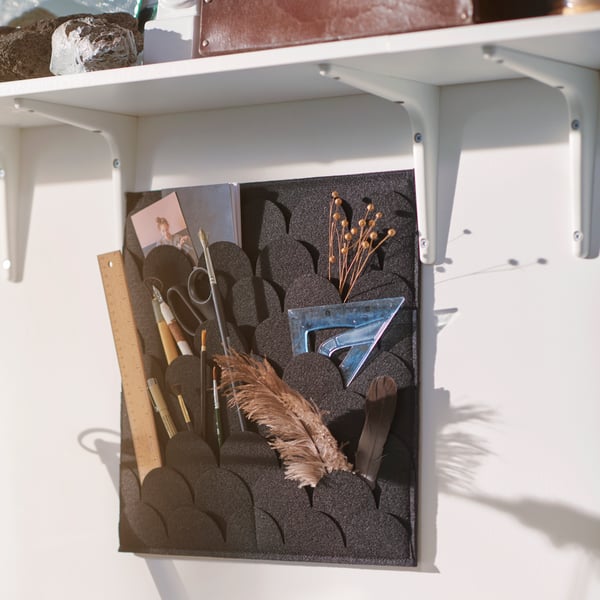 A PLUGGLAND memo board hangs on the wall underneath a shelf. It holds pens, scissors, dried flowers and two feathers.