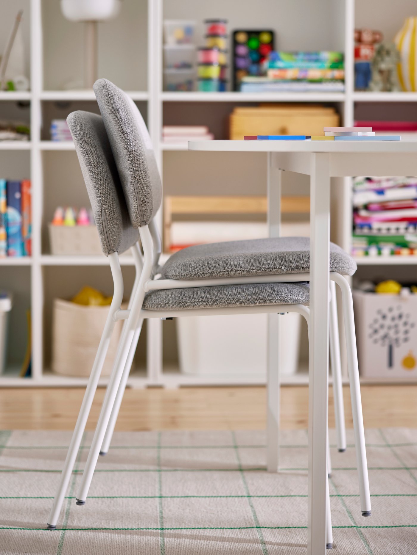 Zwei graue, gepolsterte VIHALS Stapelstühle mit weißen Metallbeinen an einem schlichten weißen Tisch, davor ein heller Teppich mit feinem Karomuster, im Hintergrund ein offenes Regal mit bunten Kisten und Spielsachen.