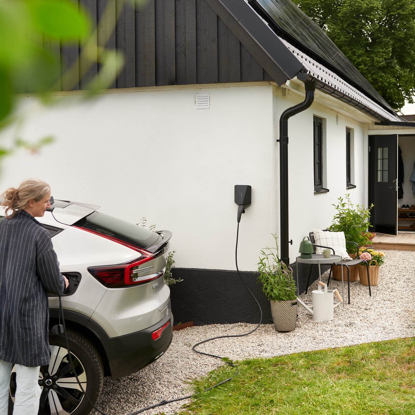 Voiture électrique raccordée à une borne de recharge installée sur le mur extérieur d’une maison moderne. L’image comprend de la verdure : des plantes, des arbres, etc.