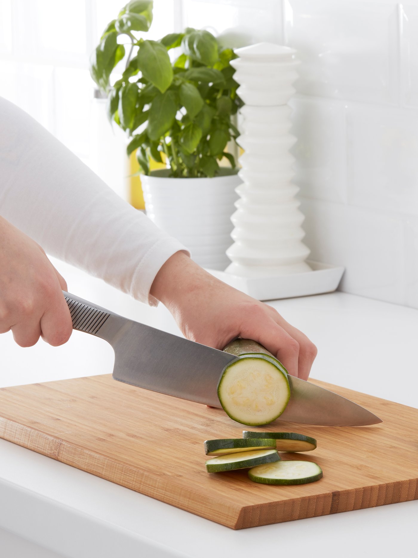 Hænderne på en person, der bruger et APTITLIG bambusskærebræt og en stor køkkenkniv til at skære en zucchini i skiver.