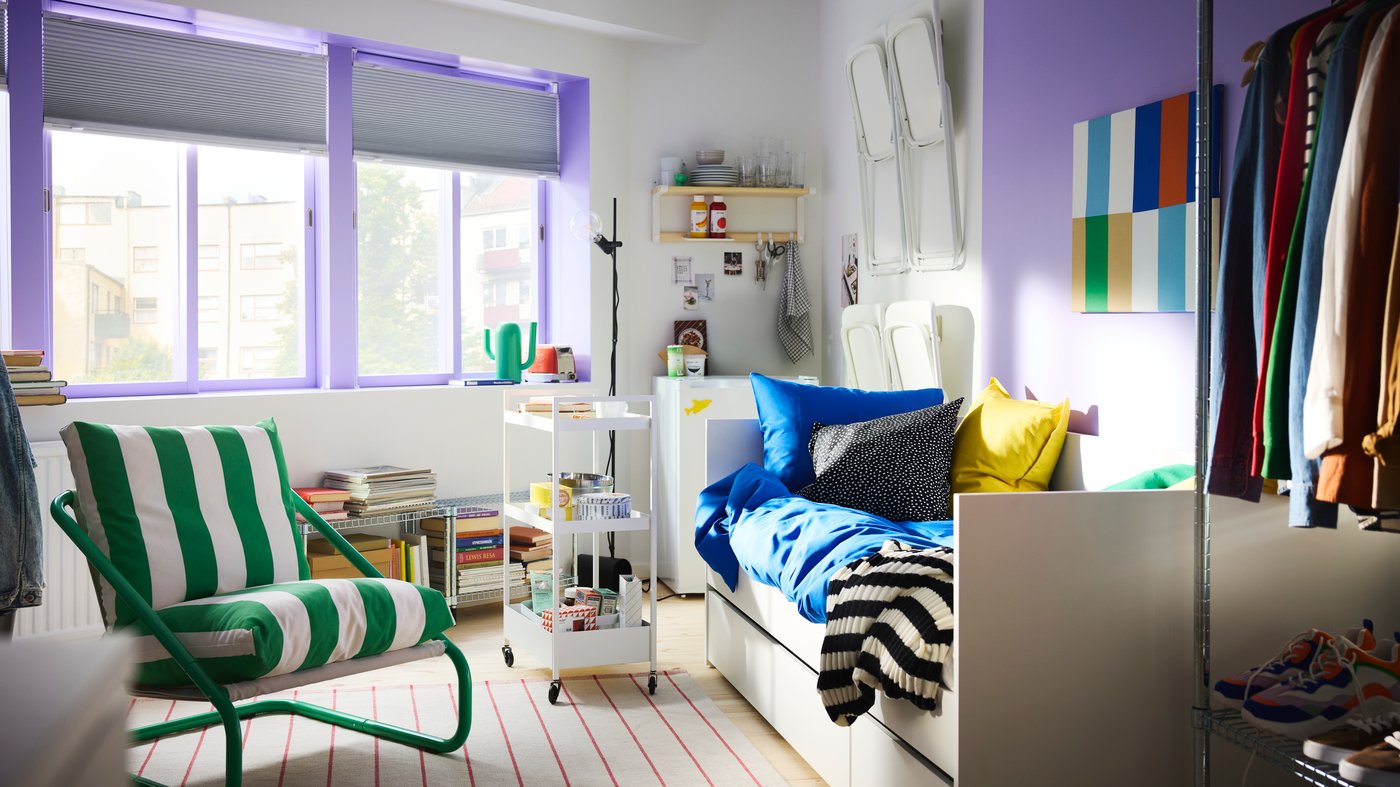 Chambre à coucher avec grandes fenêtres peintes en violet, lit banquette blanc, desserte à roulettes et fauteuil ÖNNESTAD vert/blanc.