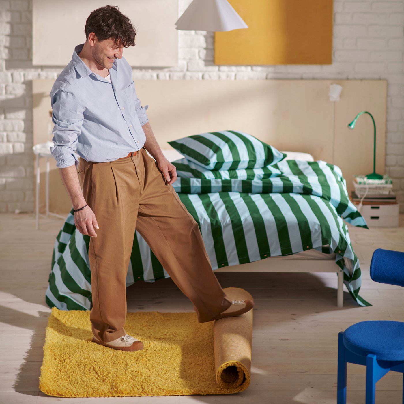 Un hombre desenrolla una alfombra ÄRENDE amarilla en un dormitorio con una cama de madera y ropa de cama SLÅNHÖSTMAL de rayas azules y verdes.