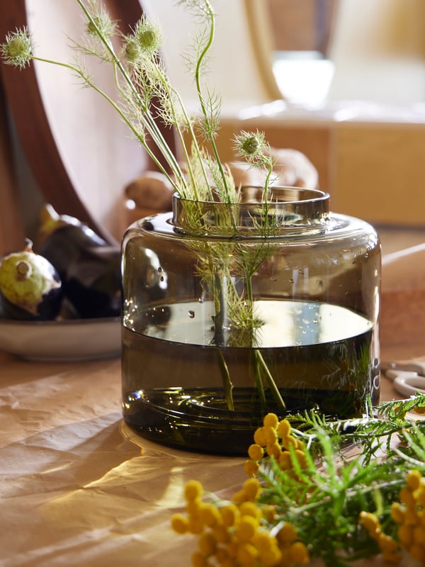 A table with a KONSTFULL vase in green-brown containing water and some green sprigs, greenery on the table, and aubergines.