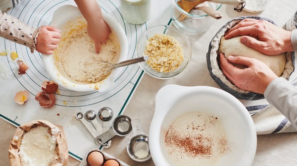 A collection of mixing bowls filled with various ingreidents as a child and adult bake together. Also are some measuring cups. 