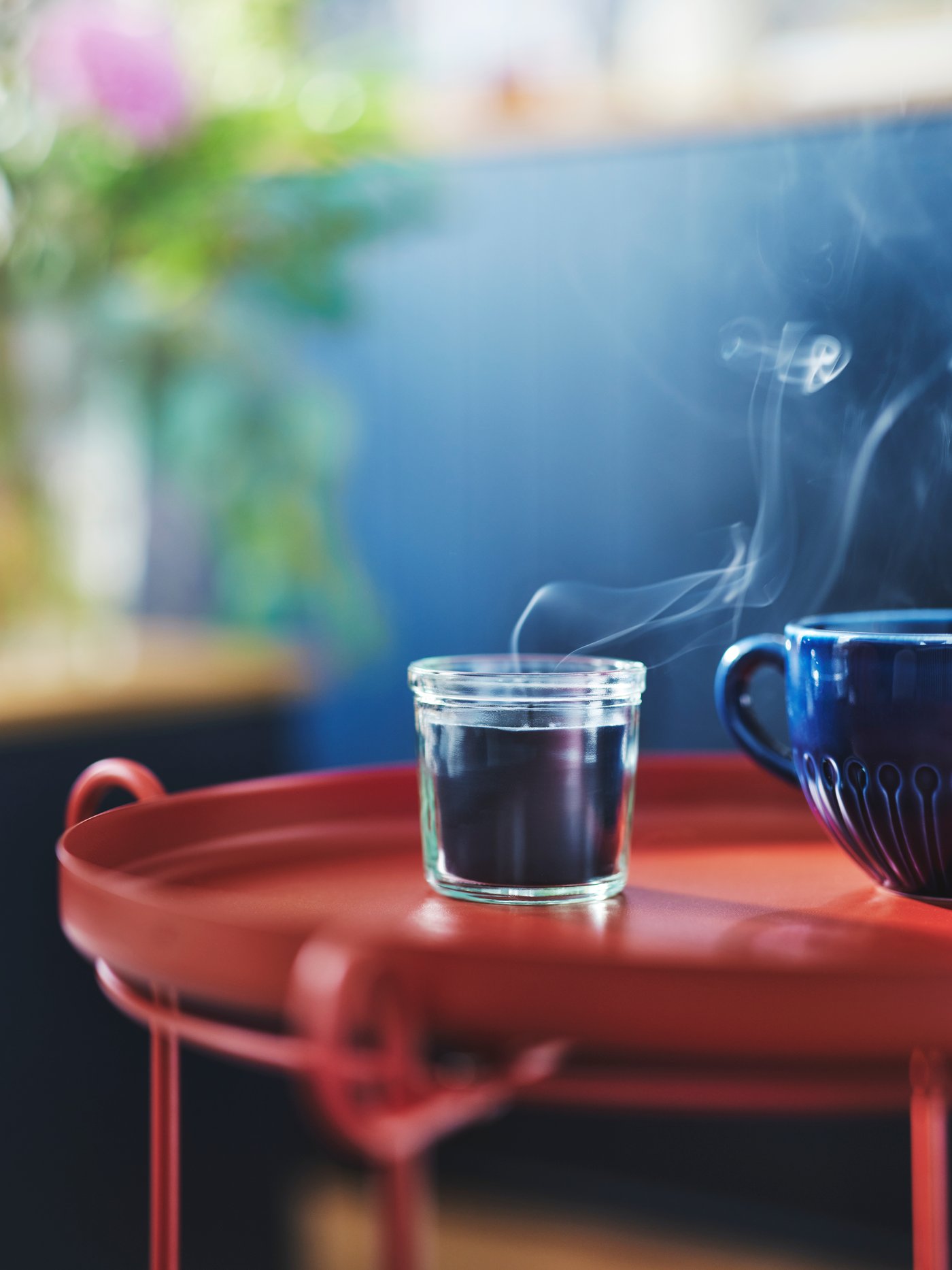 A FRUKTSKOG scented candle in glass stands next to a blue stoneware STRIMMIG mug on a red LIVELYCKE tray table.