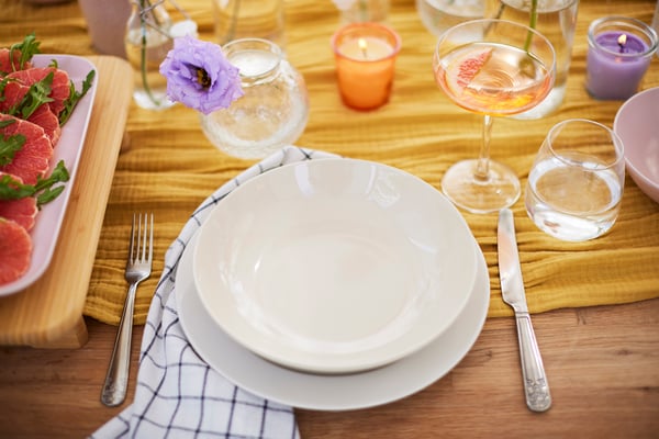 A table setting consisting of a shallow and deep plate, a champagne bowl, water glass and elegant cutlery with relief. On the table with table runner in the middle are vases with flowers and scented candles in a glass