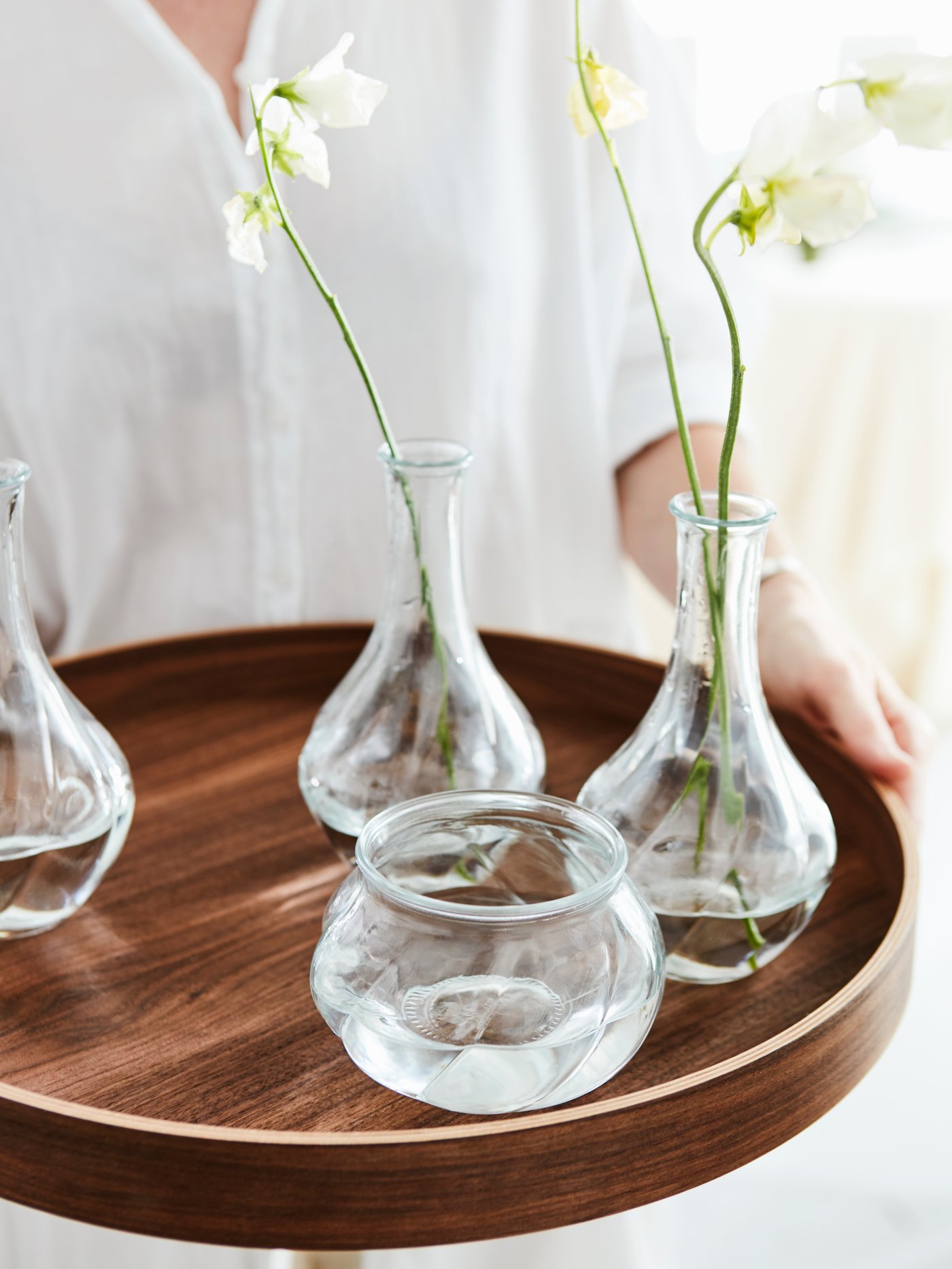En person klädd i vitt håller i en OMBONAD bricka i mörkbrunt trä med glasvaser i olika former på, några med vita blommor.