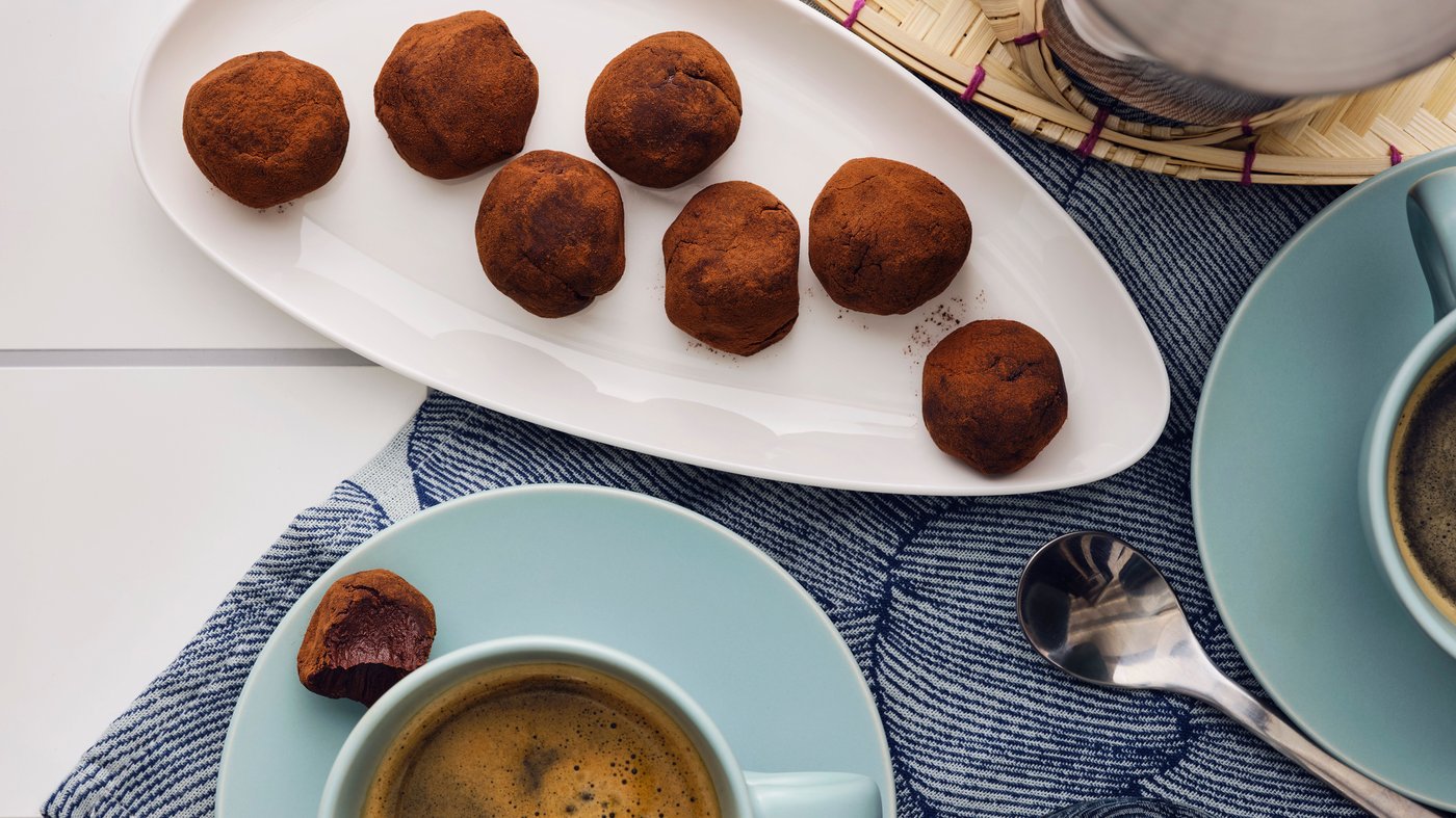 A white leaf-shaped plate with seven chocolate truffles, placed next to two turquoise cups on saucers with coffee.
