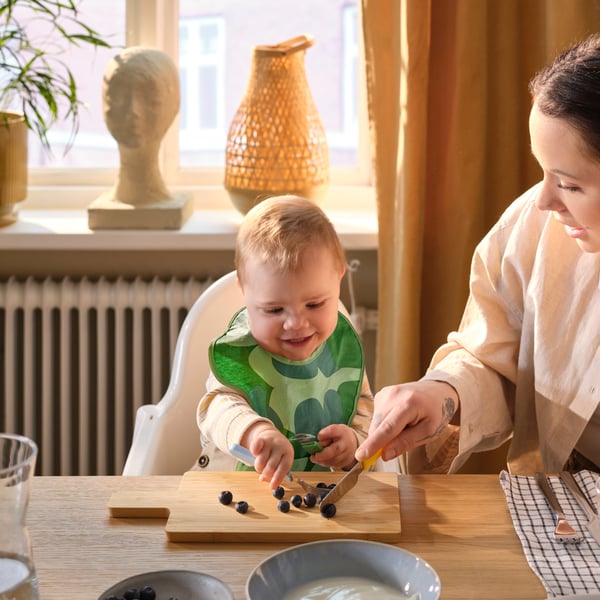Muni d’un bavoir GRÖNFINK, un enfant en bas âge assis à table mange des fruits mous sur une planche à découper à l’aide de couverts SMASKA.