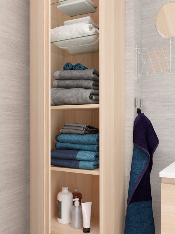 High cabinet in white stained oak effect, open to reveal lots of towels, shower products on glass and wooden shelves.