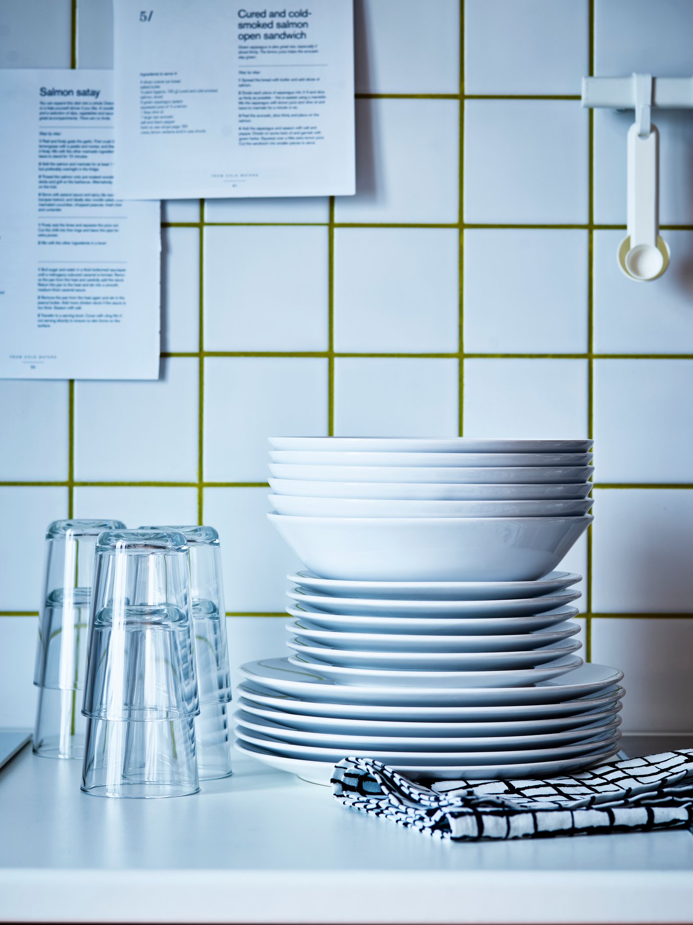 Un service de vaisselle VÄRDERA sur un comptoir blanc près de verres VARDAGEN. Il y a des carreaux blancs en arrière-plan.