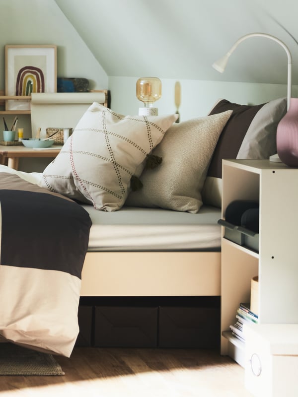 Lit GURSKEN beige clair agrémenté de divers coussins près d’une petite étagère GURSKEN où sont posés une lampe de table blanche et un vase rose.