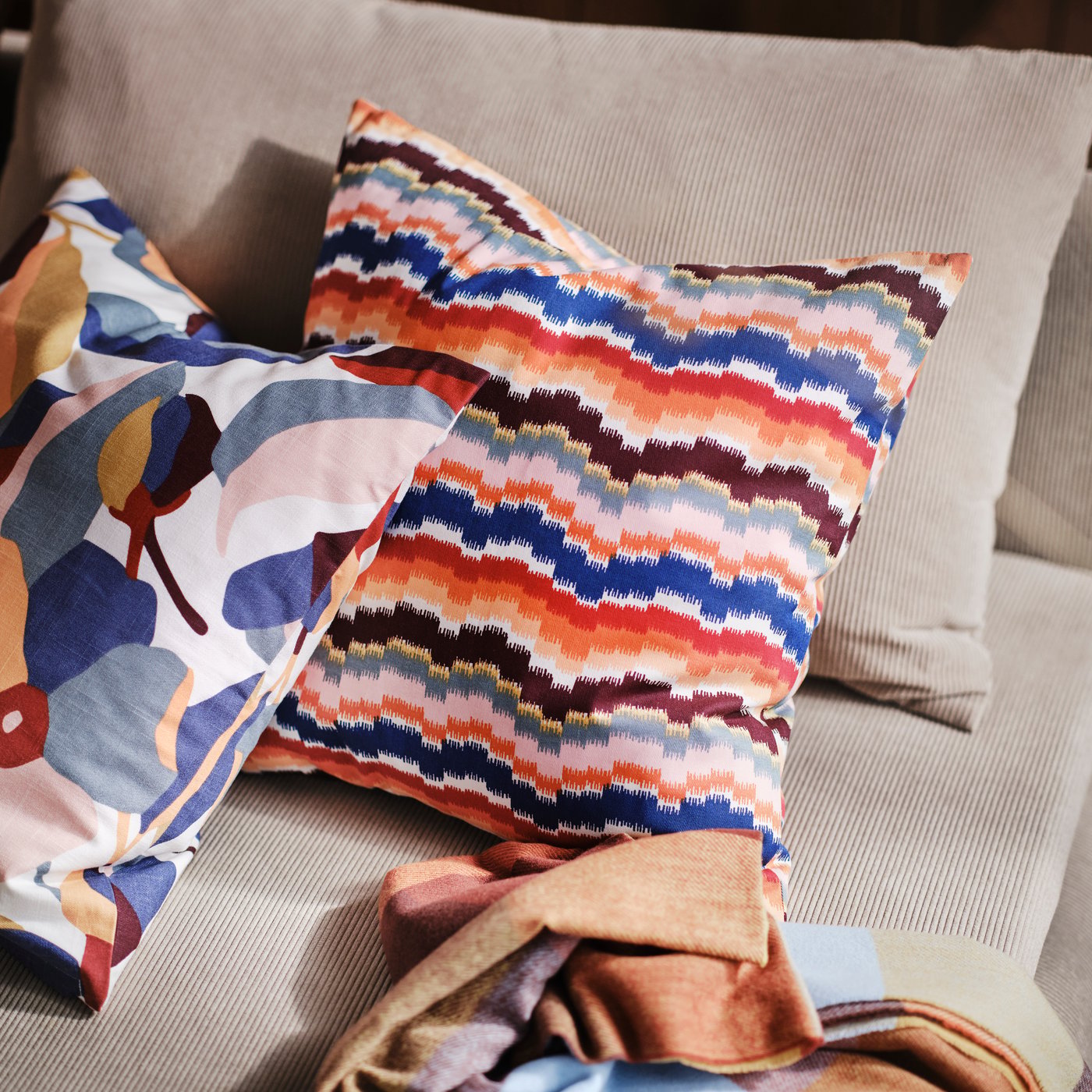 Gros plan sur deux coussins posés sur un canapé beige. L'un d'eux est doté d'une housse de coussin multicolore PEKINGSYREN à motif zigzag.