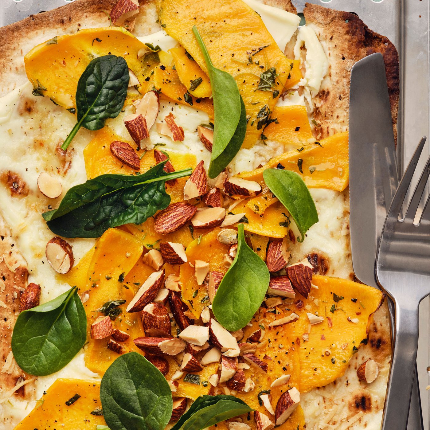 Pizza med GRÄDDAT blødt tyndt fladbrød og ristet græskar, toppet med spinat, og MUNSBIT ristede mandler, med bestik.