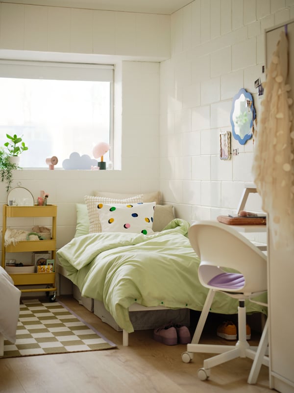 Shared dorm room with white walls showing bed with light green bedding and spotty cushion and yellow storage unit on wheels.