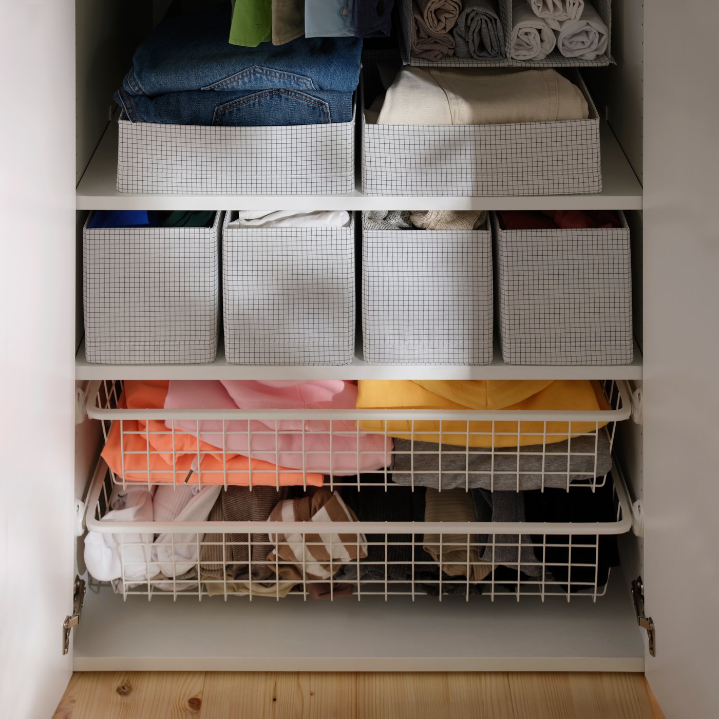 Ein offener Kleiderschrank mit gefalteter Kleidung, die in STUK‑Boxen mit Fächern auf Regalen und in Drahtschubladen verstaut ist.