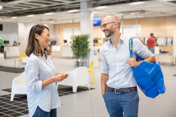 Zwei Personen unterhalten sich in einem Gang im IKEA Einrichtungshaus. EIne Person hat eine blaue IKEA Tasche über der Schulter hängen.